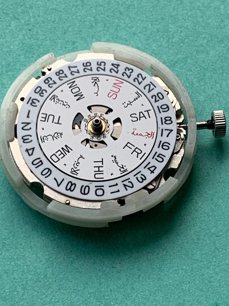 May include: A close-up of a watch movement with a white dial and silver-colored components. The dial displays the days of the week in English and Arabic, along with the date. A small crown is visible on the right side.
