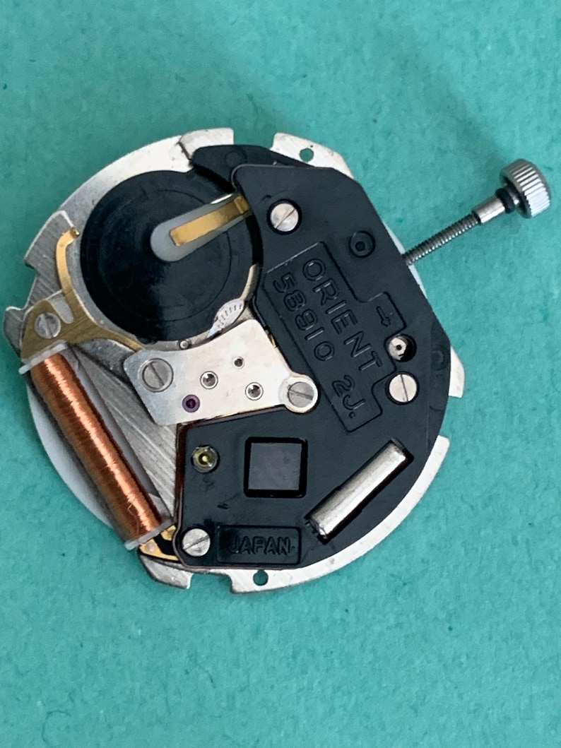 May include: A close-up of a watch movement with the text "ORIENT 58910 2J" and "JAPAN". The movement features silver, black, and copper components, including a small screw and a coil. The watch mechanism is circular.