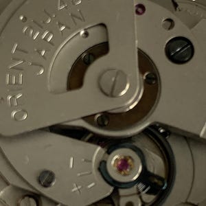 May include: Close-up of a watch movement with the text "ORIENT JAPAN" and "21J 46943". The mechanism features silver-colored metal components, a ruby jewel, and various gears and screws. The image highlights the intricate details of the watch's internal workings.