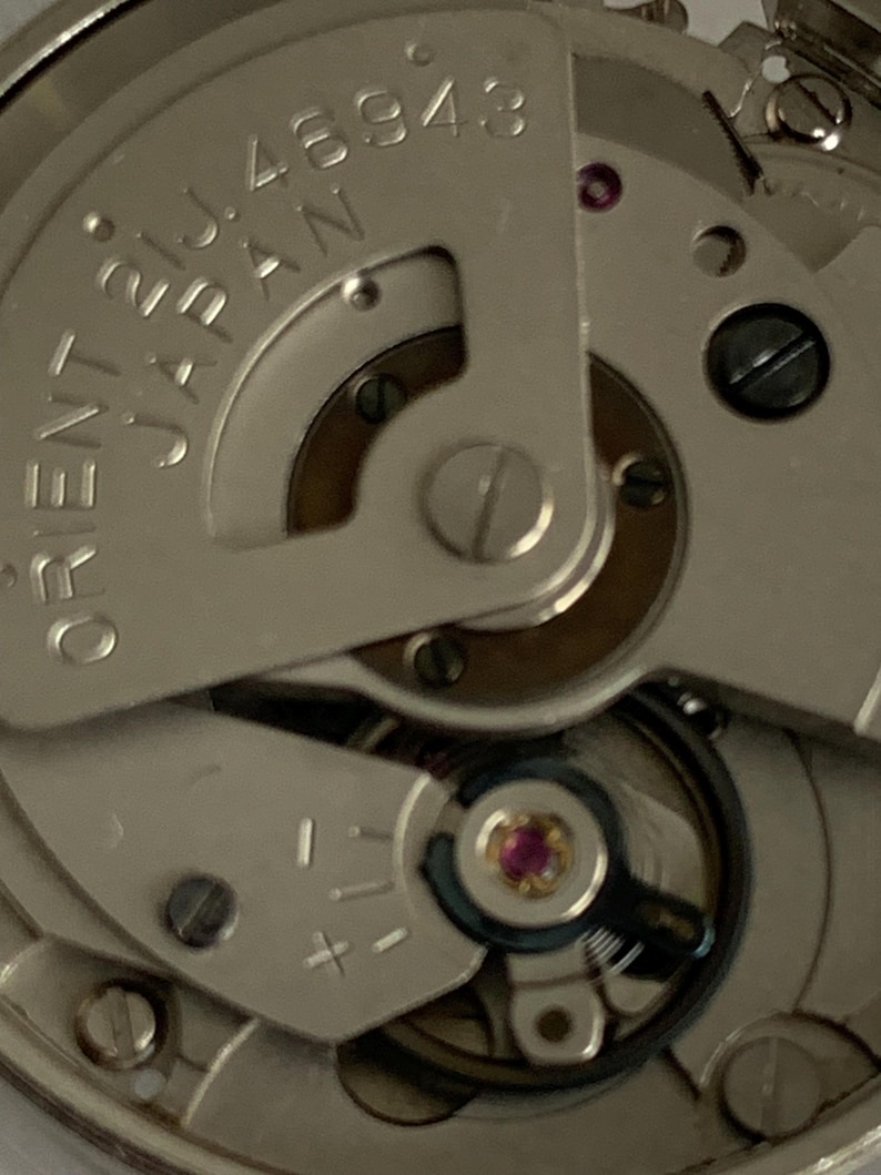 May include: Close-up of a watch movement with the text "ORIENT JAPAN" and "21J 46943". The mechanism features silver-colored metal components, a ruby jewel, and various gears and screws. The image highlights the intricate details of the watch's internal workings.