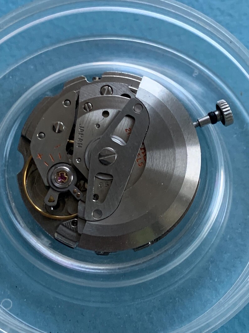 May include: A detailed view of a watch movement, featuring silver-toned gears, springs, and a balance wheel. The word "JAPAN" is stamped on a component. The mechanism is housed within a clear, circular container.