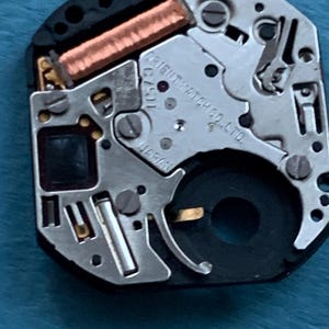 May include: Close-up of a watch movement with silver and black components. The text "ORIENT WATCH CO. LTD." and "JAPAN" are visible. A copper-colored coil and small screws are also present.