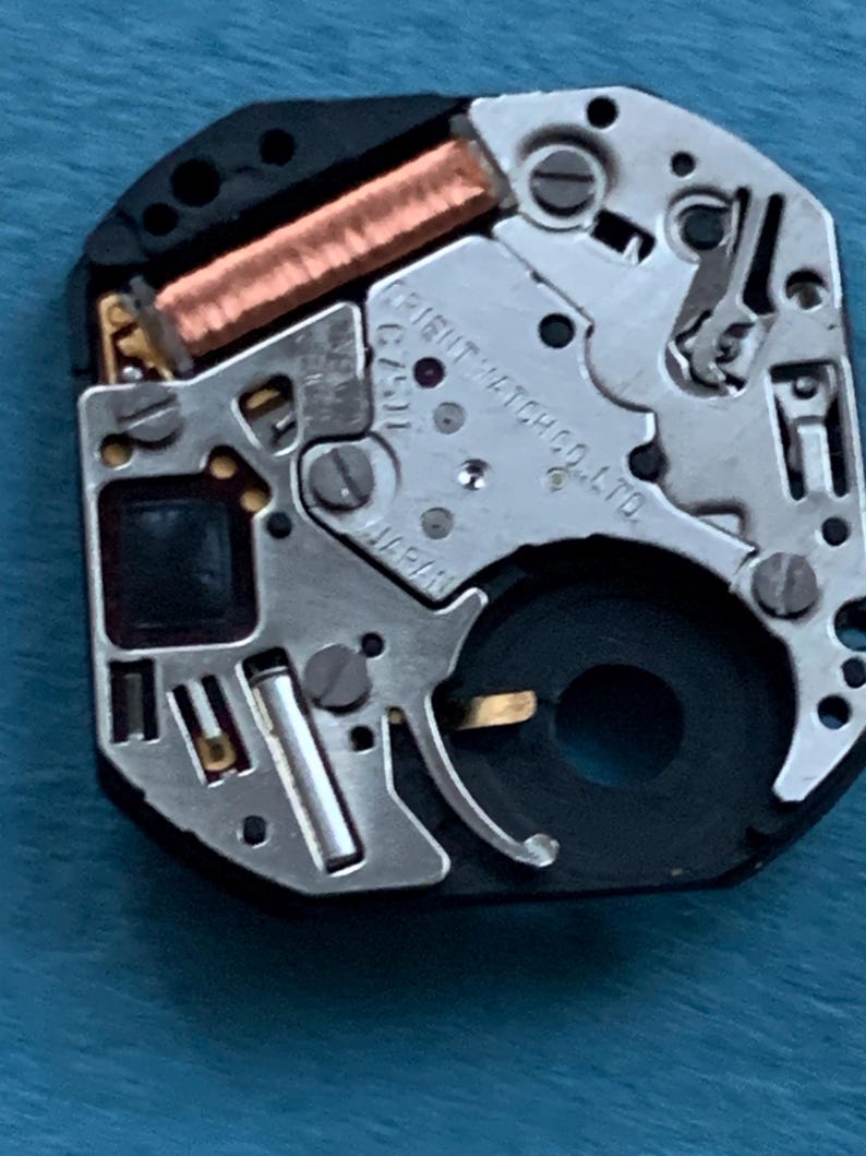 May include: Close-up of a watch movement with silver and black components. The text "ORIENT WATCH CO. LTD." and "JAPAN" are visible. A copper-colored coil and small screws are also present.