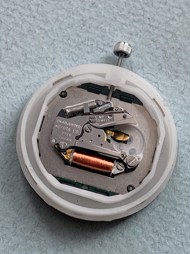 Puede incluir: Primer plano de un movimiento de reloj con el texto "UNADJUSTED MIYOTA CO. 2115 JAPAN". El movimiento presenta una bobina de cobre, engranajes plateados y una carcasa de pl&aacute;stico blanco. Se ve un tornillo plateado en la parte superior.