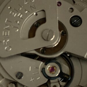 May include: Close-up of a silver watch movement with the text "ORIENT JAPAN" and "21J 46943". The watch mechanism features various gears, screws, and a jewel-toned ruby. The image highlights the intricate details of the watch's internal components.