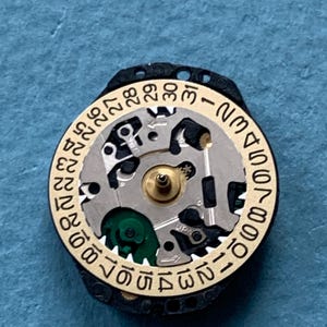 K&ouml;nnte beinhalten: Nahaufnahme eines Uhrwerks mit einem goldfarbenen Ring, der die Tage des Monats von 1 bis 31 anzeigt. Das Uhrwerk hat silberne und schwarze Komponenten, mit einem gr&uuml;nen Element. Der Uhrenmechanismus ist vor blauem Hintergrund platziert.