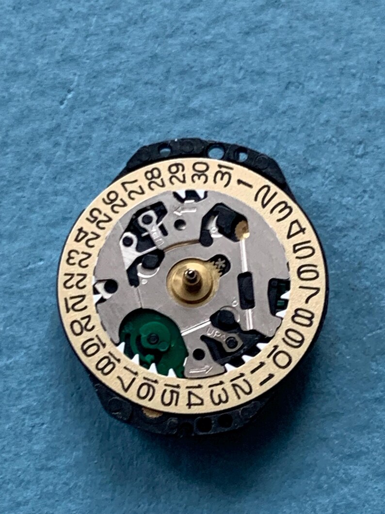 K&ouml;nnte beinhalten: Nahaufnahme eines Uhrwerks mit einem goldfarbenen Ring, der die Tage des Monats von 1 bis 31 anzeigt. Das Uhrwerk hat silberne und schwarze Komponenten, mit einem gr&uuml;nen Element. Der Uhrenmechanismus ist vor blauem Hintergrund platziert.