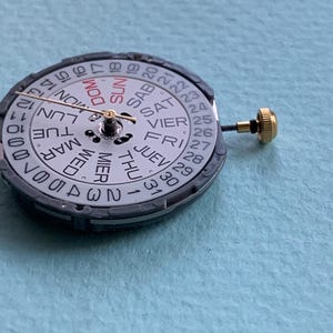 K&ouml;nnte beinhalten: Nahaufnahme eines Uhrwerks mit wei&szlig;em Zifferblatt. Das Zifferblatt zeigt die Wochentage und das Datum in Schwarz und Rot an. Eine goldfarbene Krone ist seitlich sichtbar.