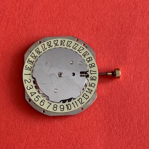 K&ouml;nnte beinhalten: Ein Uhrwerk mit einem wei&szlig;en Datumsrad, das die Zahlen 1-31 anzeigt. Das Uhrwerk hat eine silberfarbene Metallbasis, eine goldfarbene Krone und einen roten Hintergrund.