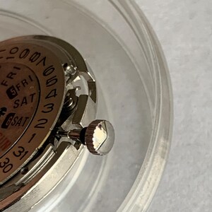 May include: Close-up of a watch movement with a silver-colored crown and a dial displaying the days of the week and dates. The watch mechanism is partially visible within a clear, circular container.