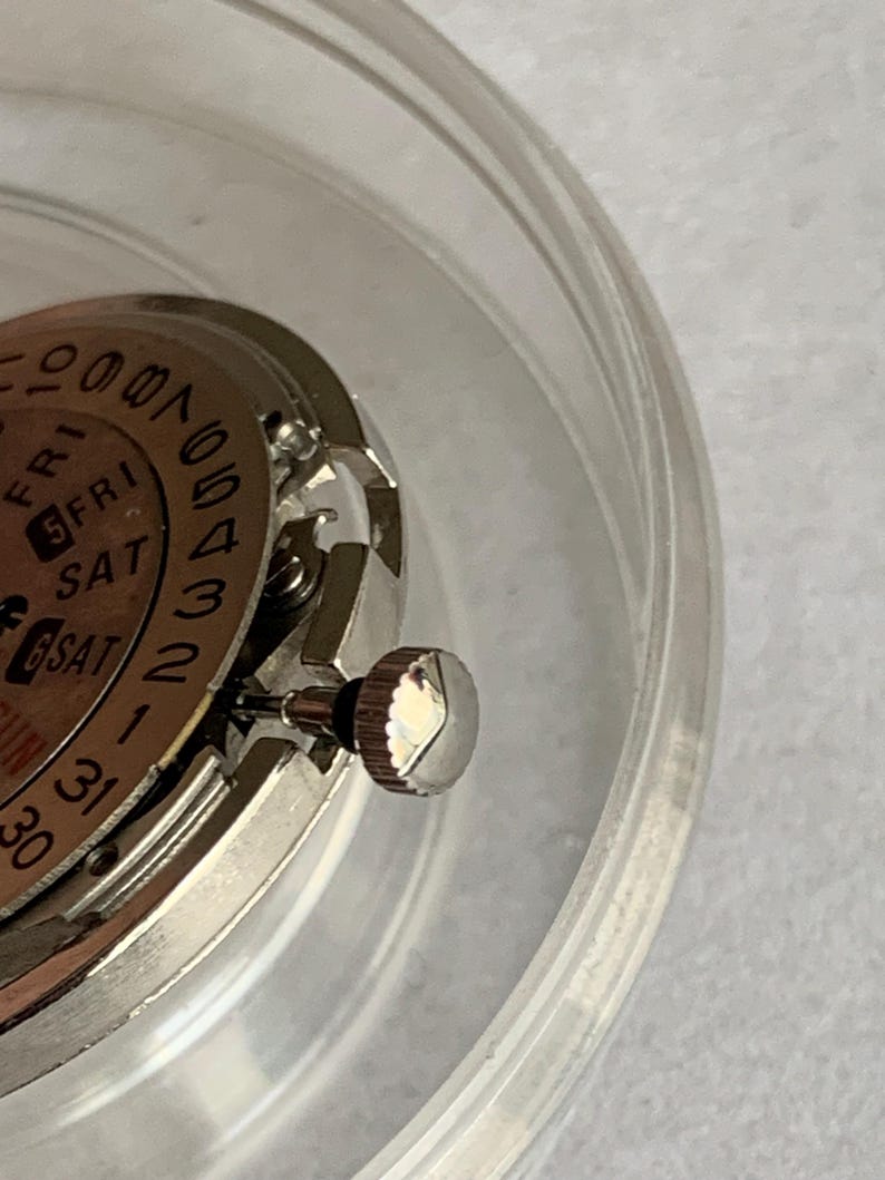 May include: Close-up of a watch movement with a silver-colored crown and a dial displaying the days of the week and dates. The watch mechanism is partially visible within a clear, circular container.