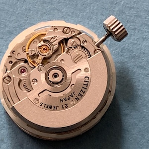 May include: Close-up of a Citizen watch movement with intricate gears, jewels, and a winding stem. The watch mechanism is silver-toned with the text "CITIZEN JAPAN 21 JEWELS" visible. The background is a solid blue.