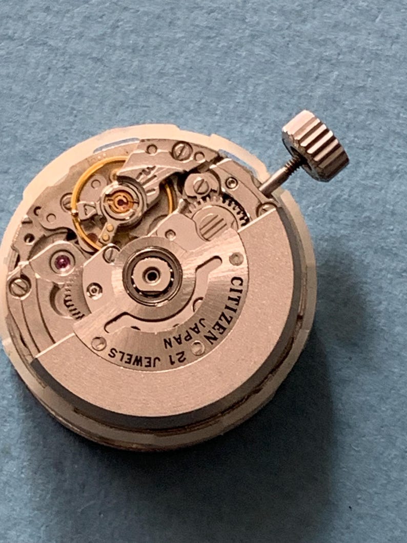 May include: Close-up of a Citizen watch movement with intricate gears, jewels, and a winding stem. The watch mechanism is silver-toned with the text "CITIZEN JAPAN 21 JEWELS" visible. The background is a solid blue.
