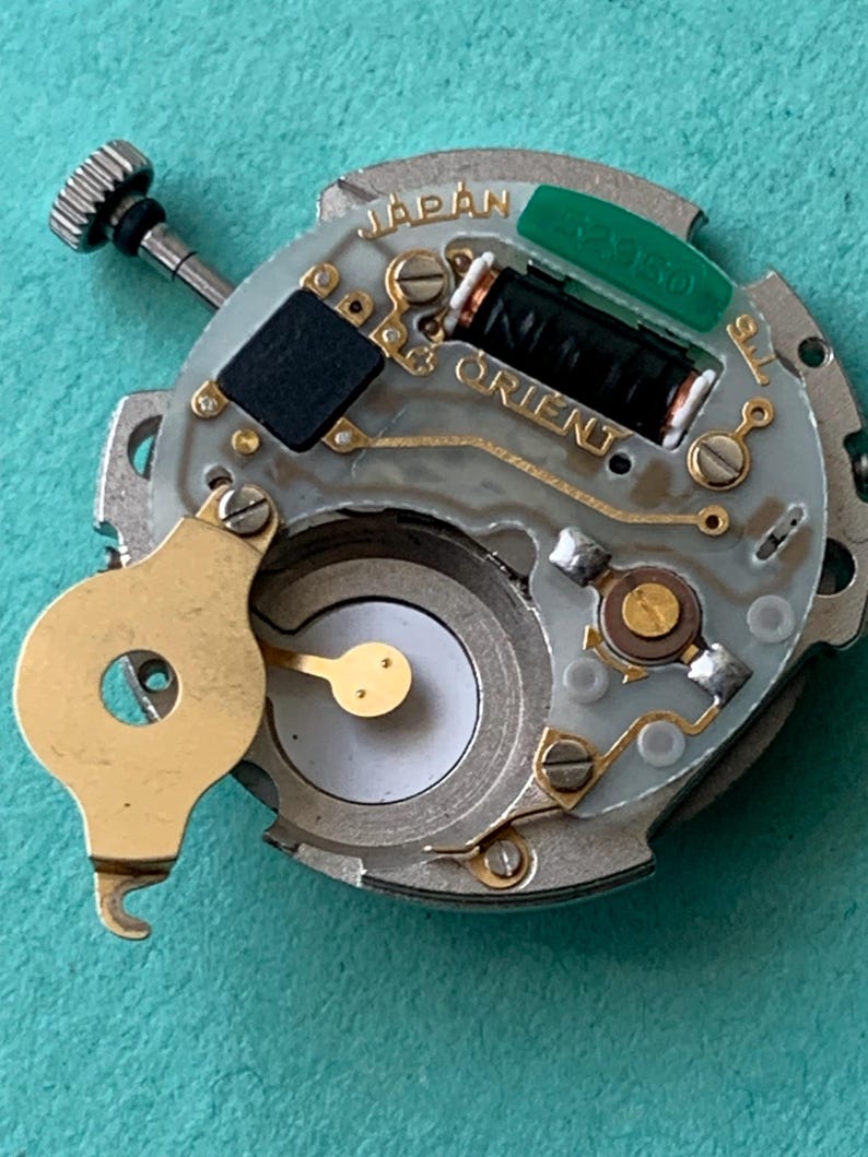 May include: A close-up of a watch movement with the word "JAPAN" and "ORIENT" visible. The movement has a silver base with gold-colored components, a black rectangular piece, and a green component. A small, silver-colored screw is visible.