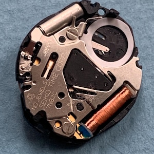 K&ouml;nnte beinhalten: Nahaufnahme eines Uhrwerks mit komplizierten mechanischen Komponenten. Das Uhrwerk verf&uuml;gt &uuml;ber metallische Silber- und Goldteile, eine Kupferspule und den Text "NO JEWELS SEIKO TIME CORP. 7N82C JAPAN". Das Uhrwerk befindet sich auf einer blauen Oberfl&auml;che.