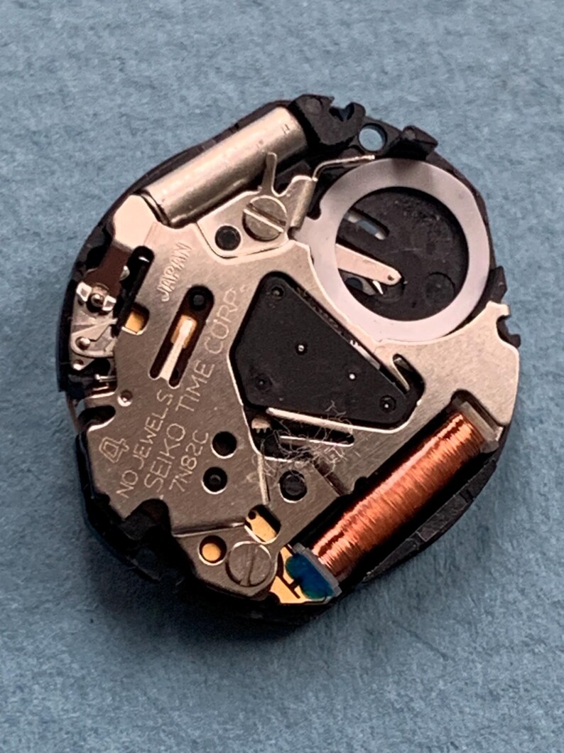 K&ouml;nnte beinhalten: Nahaufnahme eines Uhrwerks mit komplizierten mechanischen Komponenten. Das Uhrwerk verf&uuml;gt &uuml;ber metallische Silber- und Goldteile, eine Kupferspule und den Text "NO JEWELS SEIKO TIME CORP. 7N82C JAPAN". Das Uhrwerk befindet sich auf einer blauen Oberfl&auml;che.
