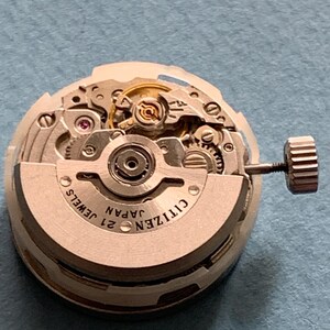 May include: A close-up of a Citizen watch movement with intricate gears and components. The watch mechanism is silver-toned with the text "CITIZEN JAPAN 21 JEWELS" visible. A small crown is attached to the side. The background is a light blue surface.