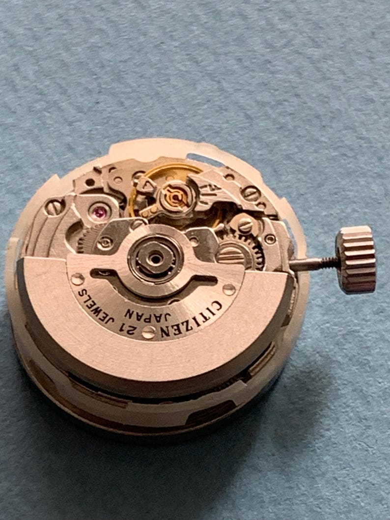 May include: A close-up of a Citizen watch movement with intricate gears and components. The watch mechanism is silver-toned with the text "CITIZEN JAPAN 21 JEWELS" visible. A small crown is attached to the side. The background is a light blue surface.