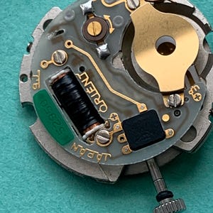 May include: Close-up of a watch movement with the word "ORIENT" visible. The movement has a gray and gold color scheme with a black coil and a green component labeled "52950". The text "JAPAN" is also visible.