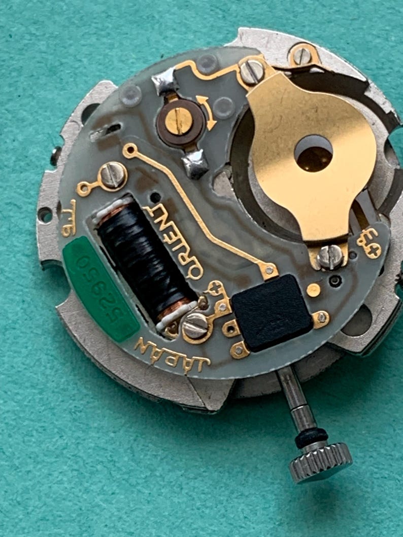 May include: Close-up of a watch movement with the word "ORIENT" visible. The movement has a gray and gold color scheme with a black coil and a green component labeled "52950". The text "JAPAN" is also visible.