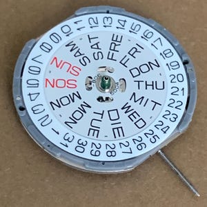 May include: A watch movement component with a white dial featuring the days of the week in English, German, and abbreviations. The dial also displays the numbers 1-31 for the date. A small metal pin extends from the side.