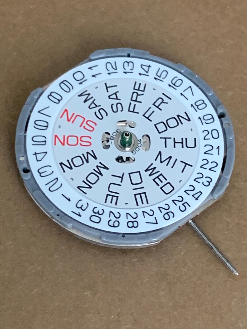 May include: A watch movement component with a white dial featuring the days of the week in English, German, and abbreviations. The dial also displays the numbers 1-31 for the date. A small metal pin extends from the side.