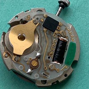 May include: Close-up of a watch movement with the word "ORIENT" visible. The movement features a silver-colored base with gold-toned components, a black coil, and a green component labeled "JAPAN". A small, textured crown is at the top.