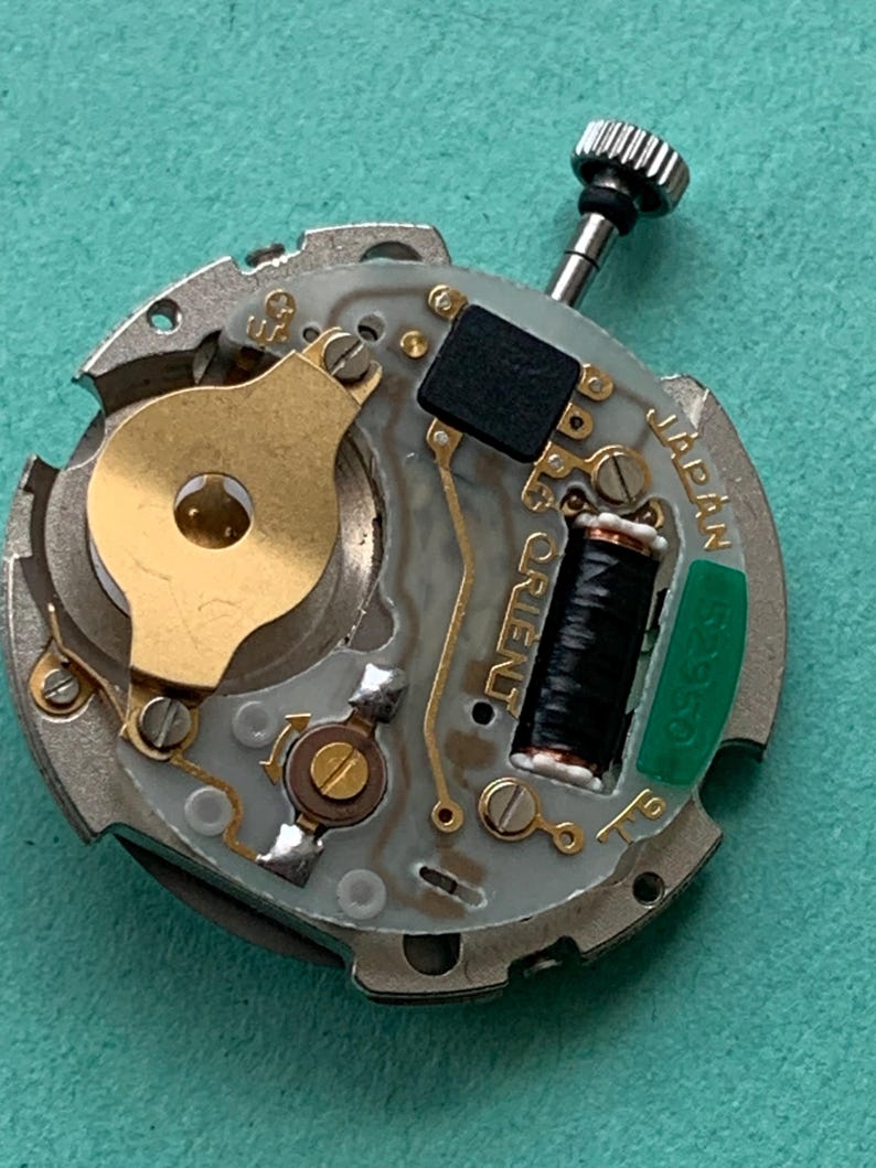 May include: Close-up of a watch movement with the word "ORIENT" visible. The movement features a silver-colored base with gold-toned components, a black coil, and a green component labeled "JAPAN". A small, textured crown is at the top.