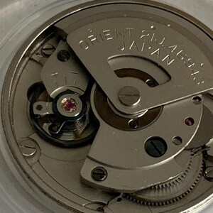 May include: Close-up of a watch movement with the text "ORIENT 21J JAPAN 46943" on a silver-colored plate. The intricate mechanism features gears, screws, and a small red jewel. The watch movement is encased in a clear, circular container.