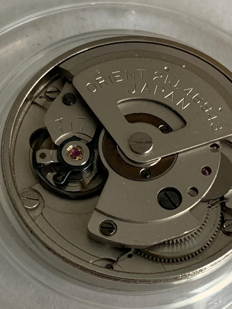 May include: Close-up of a watch movement with the text "ORIENT 21J JAPAN 46943" on a silver-colored plate. The intricate mechanism features gears, screws, and a small red jewel. The watch movement is encased in a clear, circular container.