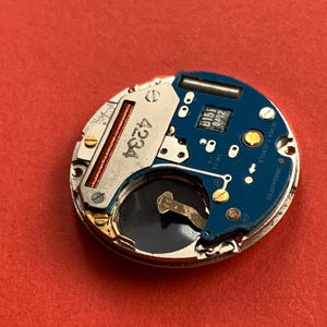 May include: Close-up of a watch movement with a blue circuit board and silver metal components. The watch movement has the number 4234 printed on it. The words "SWISS" and "SEVEN 7 JEWELS" are also visible.