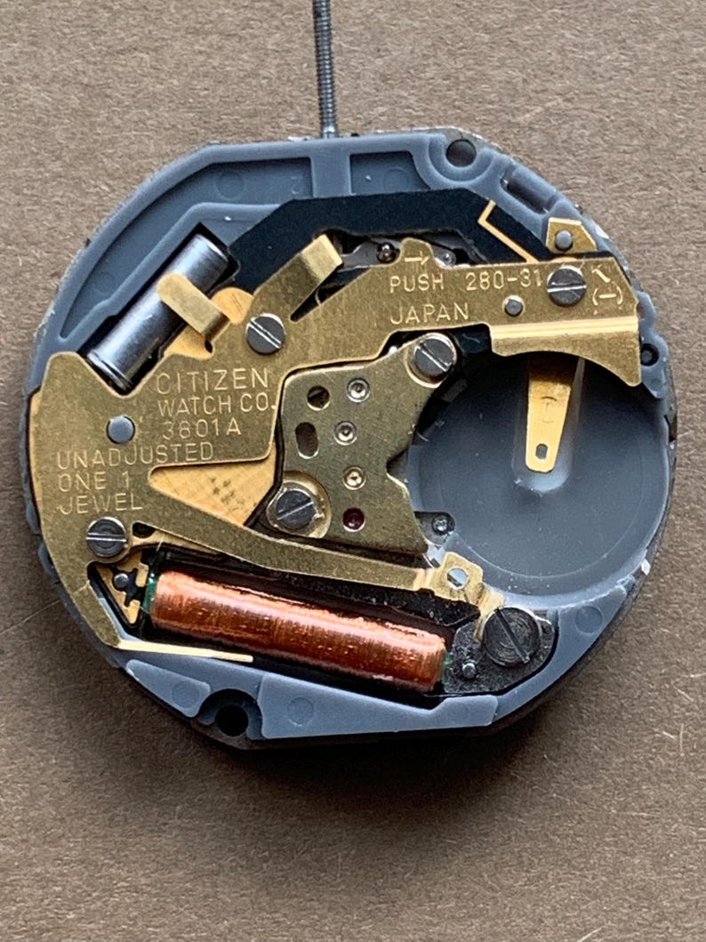 May include: A close-up of a Citizen watch movement, model 3601A, with gold-colored gears and a copper-colored coil. The watch mechanism is gray and has the text "PUSH 280-31 JAPAN" and "ONE 1 JEWEL".