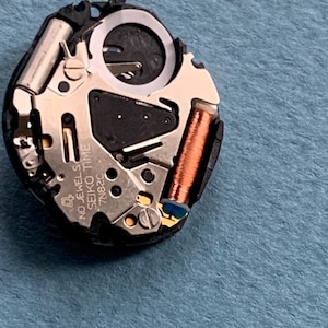 K&ouml;nnte beinhalten: Nahaufnahme eines Seiko Time 7N82C Uhrwerks. Der Uhrenmechanismus besteht aus silbernen, kupferfarbenen und schwarzen Komponenten, darunter eine Spule, Zahnr&auml;der und ein kreisf&ouml;rmiger Ring. Der Text "4 NO JEWELS SEIKO TIME 7N82C" ist sichtbar.