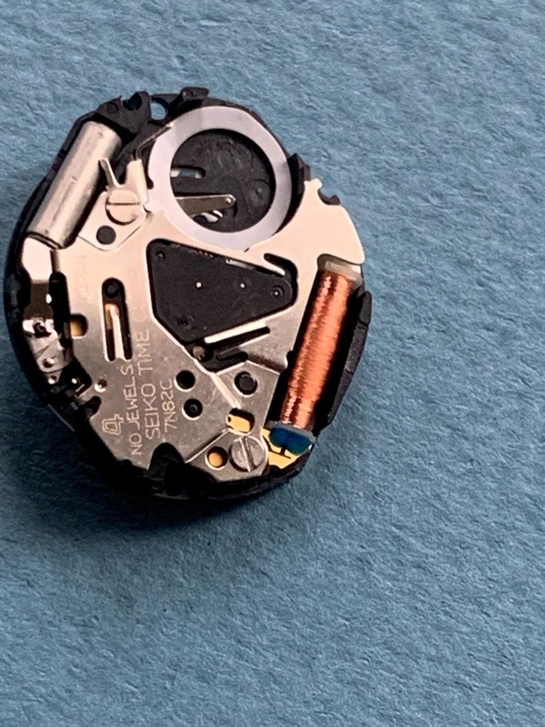 K&ouml;nnte beinhalten: Nahaufnahme eines Seiko Time 7N82C Uhrwerks. Der Uhrenmechanismus besteht aus silbernen, kupferfarbenen und schwarzen Komponenten, darunter eine Spule, Zahnr&auml;der und ein kreisf&ouml;rmiger Ring. Der Text "4 NO JEWELS SEIKO TIME 7N82C" ist sichtbar.