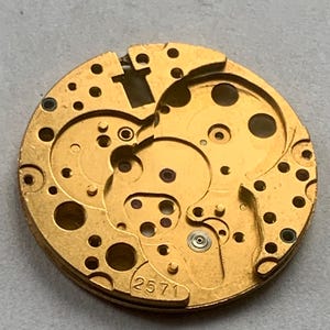 May include: Close-up of a gold-colored watch movement with intricate gears and circular cutouts. The number "2571" is visible. The watch mechanism has a complex design with multiple layers and small holes.
