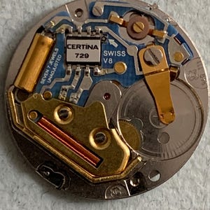 Może przedstawiać: Zbliżenie mechanizmu zegarka Certina Swiss V8. Mechanizm zegarka zawiera złote i srebrne elementy, niebieską płytkę obwodu oraz tekst "CERTINA 729" i "SWISS V8". Mechanizm zegarka to złożony układ k&oacute;ł zębatych i obwod&oacute;w.
