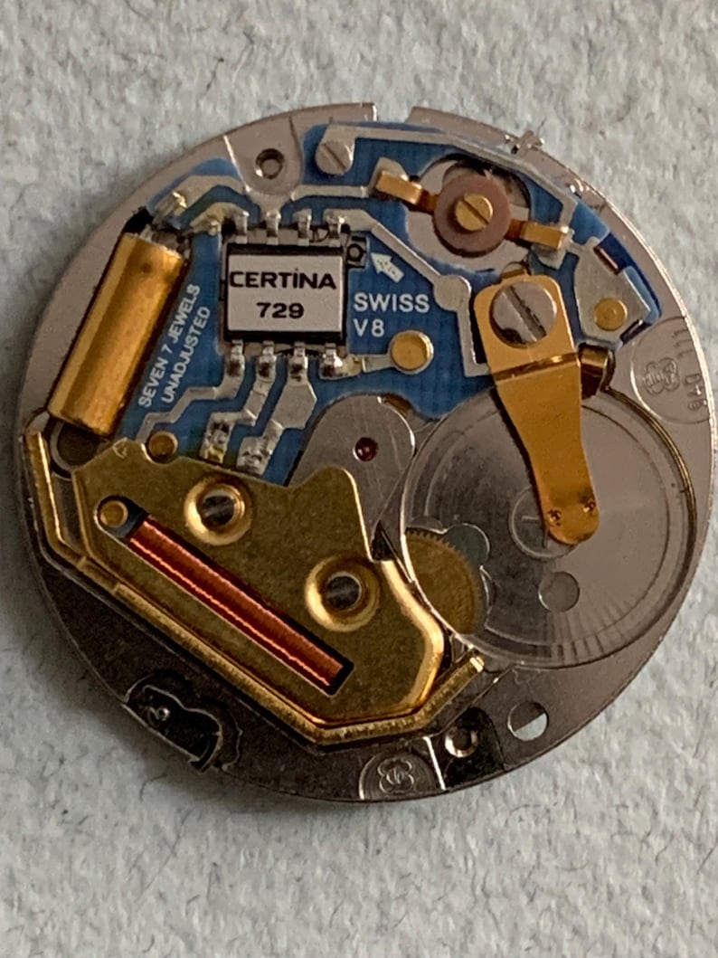 Może przedstawiać: Zbliżenie mechanizmu zegarka Certina Swiss V8. Mechanizm zegarka zawiera złote i srebrne elementy, niebieską płytkę obwodu oraz tekst "CERTINA 729" i "SWISS V8". Mechanizm zegarka to złożony układ k&oacute;ł zębatych i obwod&oacute;w.