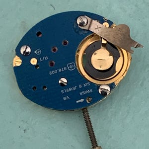 May include: Close-up of a watch movement with a blue circuit board. The watch mechanism has gold-colored components, including a circular battery holder and a screw. The text "SWISS SIX 6 JEWELS" is visible.