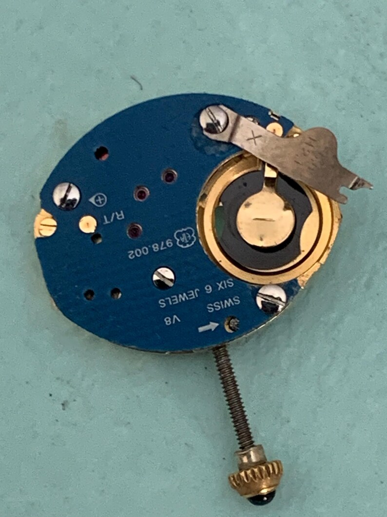 May include: Close-up of a watch movement with a blue circuit board. The watch mechanism has gold-colored components, including a circular battery holder and a screw. The text "SWISS SIX 6 JEWELS" is visible.
