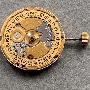 May include: Close-up of a gold-toned watch movement with intricate gears and a date dial. The dial features numbers from 1 to 31. A small screw and crown are visible on the side. The watch mechanism is set against a gray background.