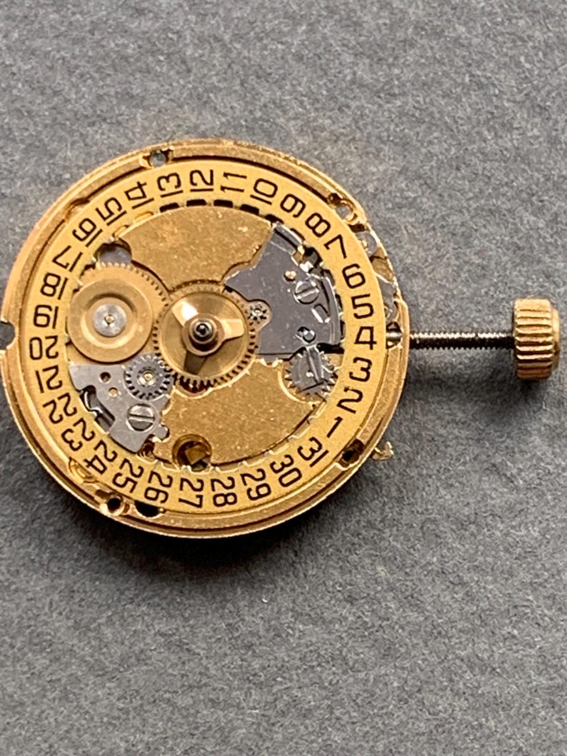 May include: Close-up of a gold-toned watch movement with intricate gears and a date dial. The dial features numbers from 1 to 31. A small screw and crown are visible on the side. The watch mechanism is set against a gray background.