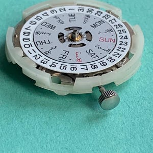 May include: Close-up of a watch movement with a white dial and a silver crown. The dial displays the days of the week in English and Arabic, along with the date. The watch mechanism is set against a teal background.