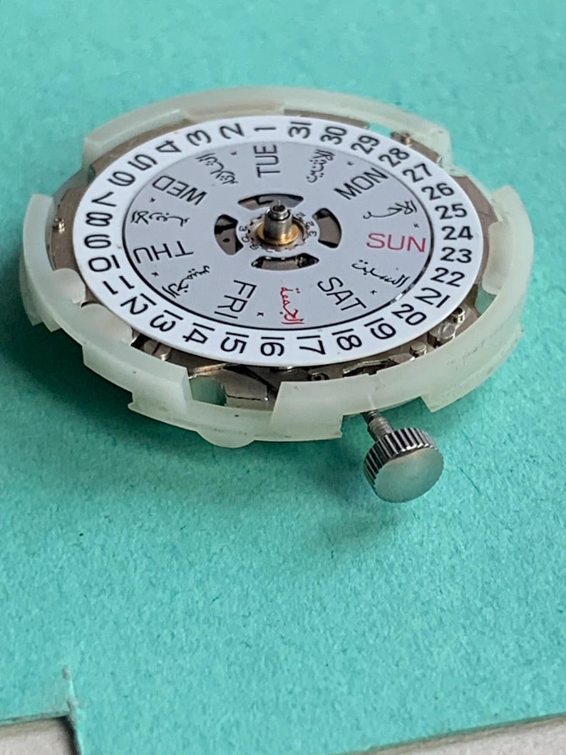 May include: Close-up of a watch movement with a white dial and a silver crown. The dial displays the days of the week in English and Arabic, along with the date. The watch mechanism is set against a teal background.