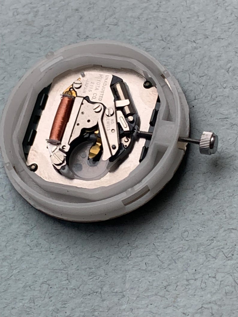 Puede incluir: Un movimiento de reloj en una carcasa de pl&aacute;stico blanco. El movimiento presenta una bobina de cobre, componentes met&aacute;licos plateados y el texto "MIYOTA CO. JAPAN 2115 UNADJUSTED NO JEWELS". Una peque&ntilde;a corona plateada est&aacute; adjunta.