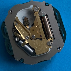 May include: Close-up of a watch movement featuring gold gears, a silver plate, and a green circuit board. The text "ONE JEWEL ADJUSTED CITIZEN WATCH CO. JAPAN" is visible. The watch mechanism is set against a blue background.