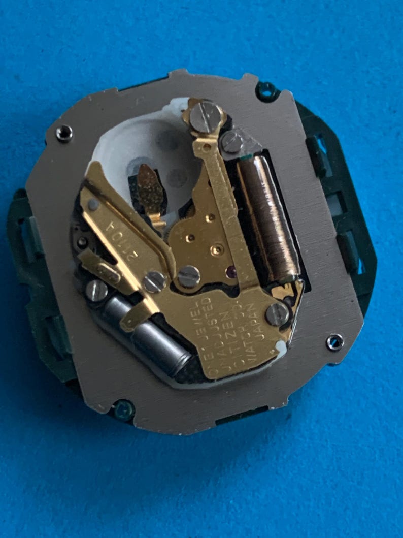 May include: Close-up of a watch movement featuring gold gears, a silver plate, and a green circuit board. The text "ONE JEWEL ADJUSTED CITIZEN WATCH CO. JAPAN" is visible. The watch mechanism is set against a blue background.