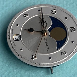 Op de afbeelding: Zilveren horlogekast met een maanfase-ontwerp. De wijzerplaat heeft zwarte wijzers, een datumvenster en het woord "QUARTZ". De wijzerplaat heeft de woorden "Swiss" en "Made" erop gedrukt.