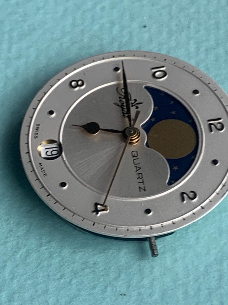 Op de afbeelding: Zilveren horlogekast met een maanfase-ontwerp. De wijzerplaat heeft zwarte wijzers, een datumvenster en het woord "QUARTZ". De wijzerplaat heeft de woorden "Swiss" en "Made" erop gedrukt.
