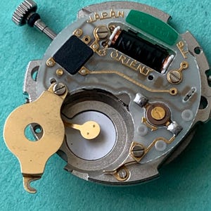 May include: A close-up of a watch movement with the word "JAPAN" and "ORIENT" visible. The movement has a silver base with gold-colored components, a black rectangular piece, and a green component. A small, silver-colored screw is visible.
