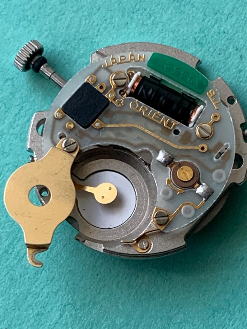 May include: A close-up of a watch movement with the word "JAPAN" and "ORIENT" visible. The movement has a silver base with gold-colored components, a black rectangular piece, and a green component. A small, silver-colored screw is visible.
